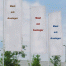 Drei Banner am 6-Meter-Fahnenmast mit drehbarem Ausleger in Braun, Rot und Blau vor bewölktem Himmel, Bäumen und Gebäuden im Hintergrund. Wertica Shop