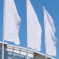 Drei leere weiße Fahnen auf 6-Meter-Masten vor Glasfassade. Produktname gelb oben rechts: "Fahnenzubehör: 6-Meter-Mast mit innenliegender Hissvorrichtung". Wertica Shop
