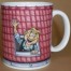 Die Dachtasse ist eine weiße Kaffeetasse mit Cartoon einer blond lächelnden Person, die aus einem Fenster in einer roten Ziegelwand winkt. Wertica Shop
