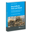 Gestapelte Dachziegel neben einer Steinmauer, blaue Akzente, fetter weißer Titel: "Baustoffe für Dach und Wand". 14. Auflage. RM Rudolf Müller. Wertica Shop